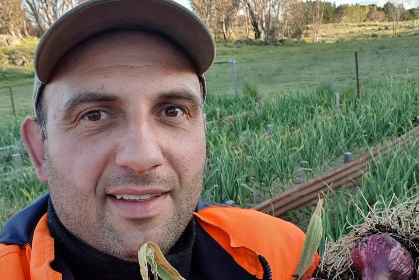 Man wearing cap and orange high-vis top smiles for camera, holding a bunch of garlic in a field.