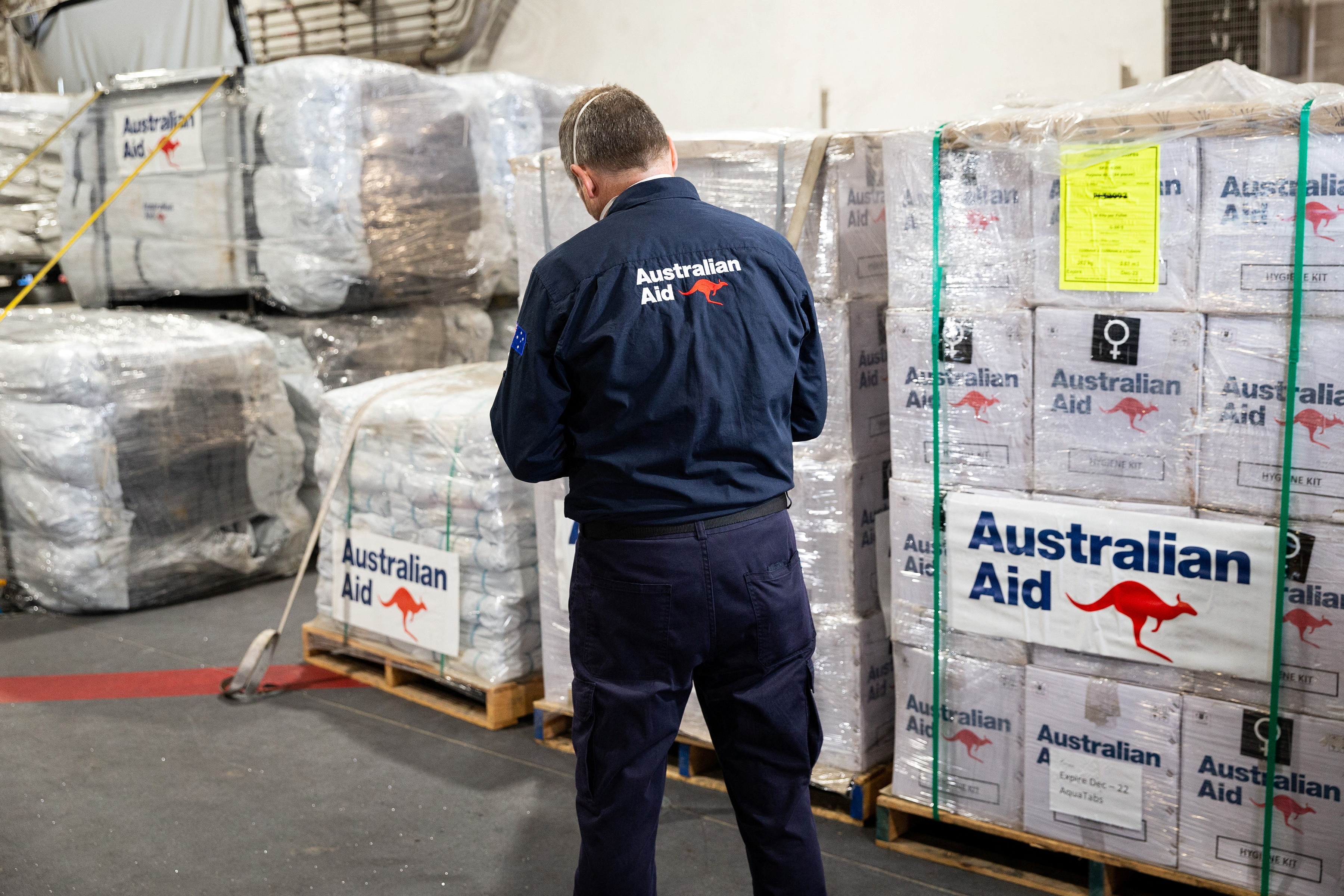 A man wearing a navy blue shirt with Australian Aid on it checks pallets of boxes.