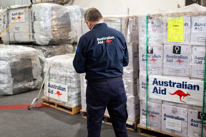 A man wearing a navy blue shirt with Australian Aid on it checks pallets of boxes.