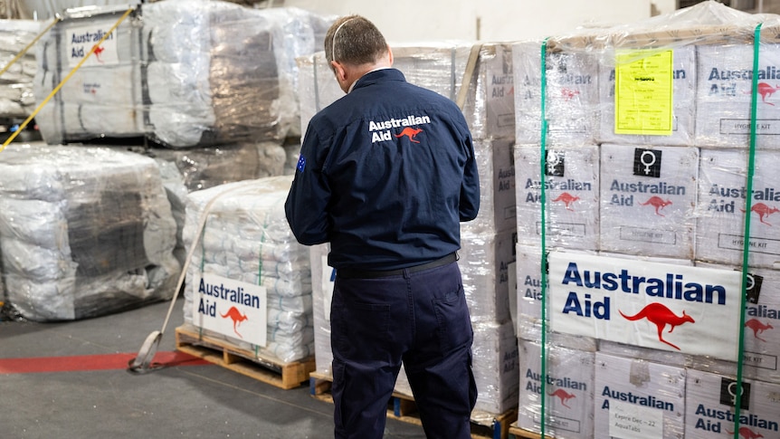A man wearing a navy blue shirt with Australian Aid on it checks pallets of boxes.