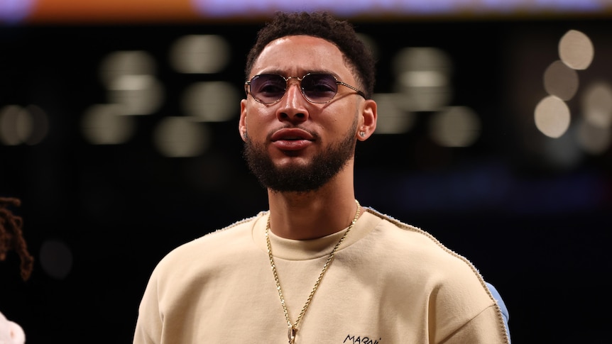 Basketballer Ben Simmons looks at the camera, wearing casual (non-playing) clothes and dark glasses during an NBA game.