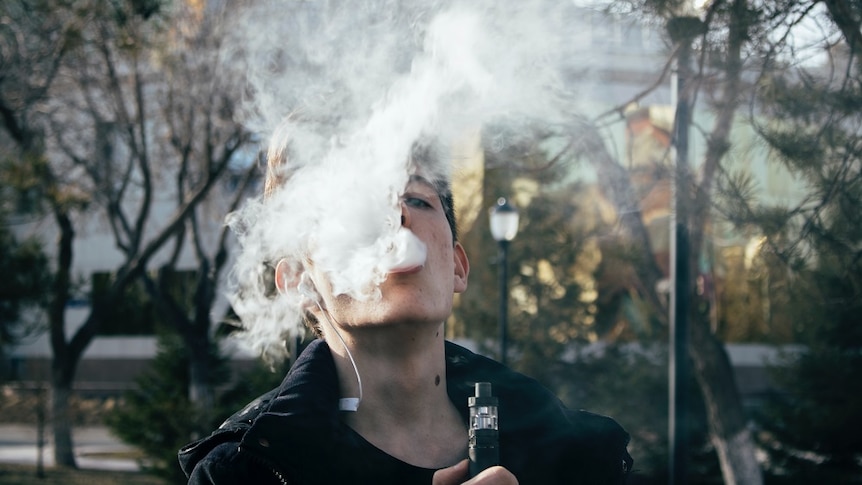 A young man in a black jacket blows out a cloud of white smoke. He holds an e-cigarette.