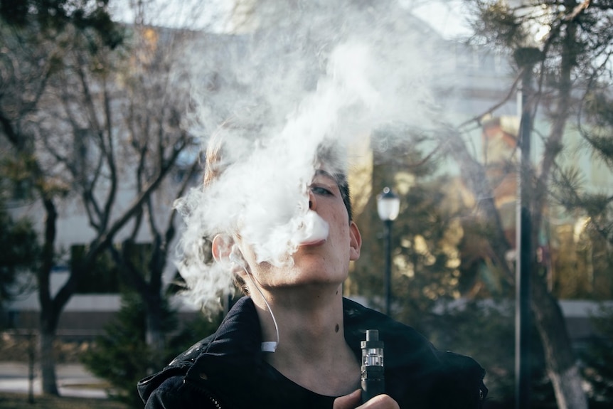 A photo of a young person vaping with their face partially obscured by the vapour cloud.