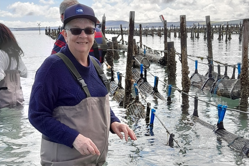 Woman in sunglasses on left looking at camera in waders in water with oyster racks in background