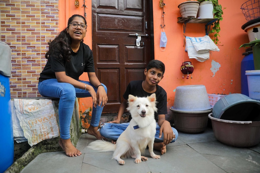 Two teenagers sit outside a house with a little white dog