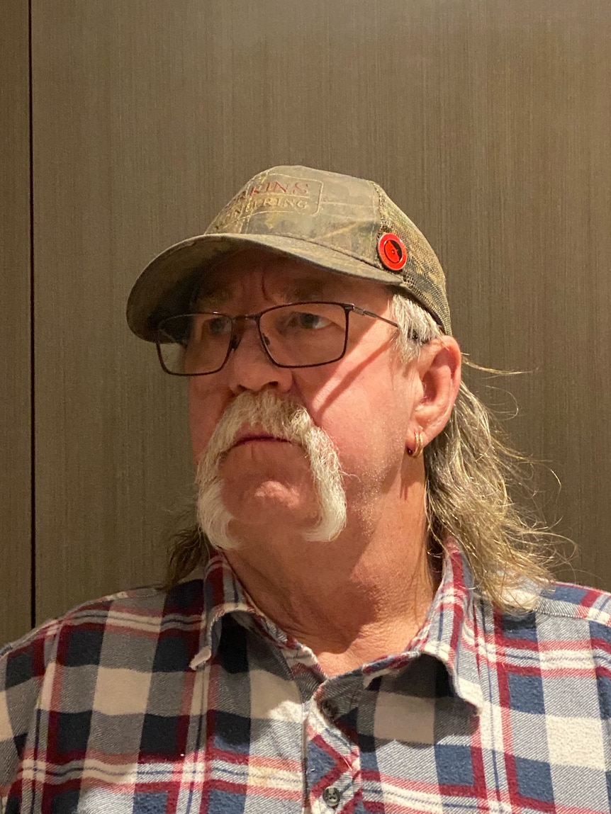 An older man with a large handlebar moustache, wearing a checked shirt and a cap.