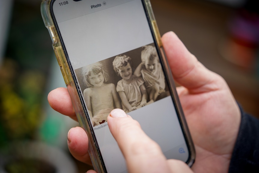 A hand holding a phone with a black and white photo of three young children on the screen. 