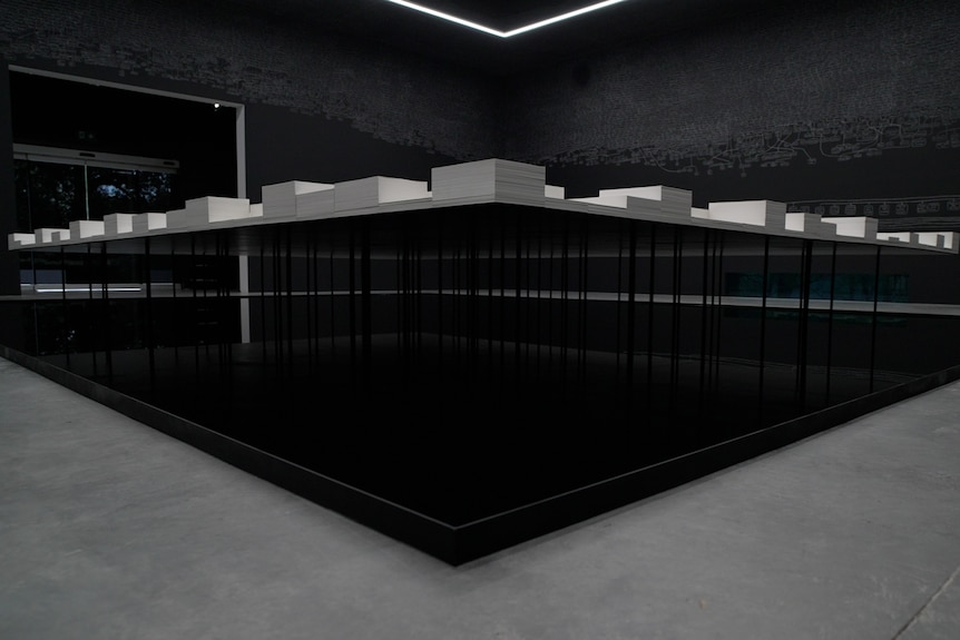 In a dimly lit gallery space a table of stacked documents stands above a shallow square pool of water above concrete floor.