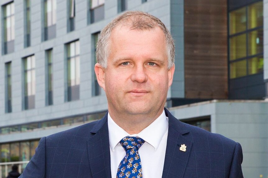 Iain Martin wearing blue suit, white shirt and blue tie, standing outside in front of a building