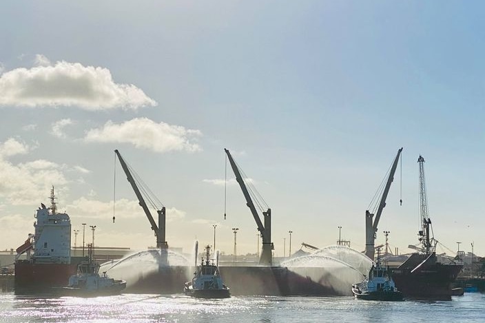 Water sprays from tug boats onto a cargo ship, with cranes in the background.