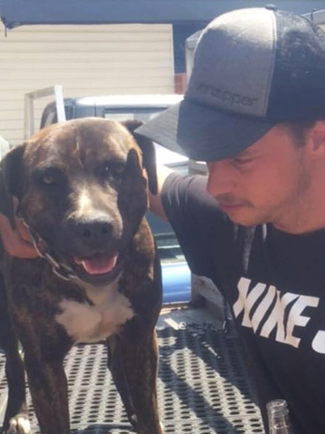 A young man wearing a black cap and shirt puts his arm around a dog, sitting on the back of a ute.