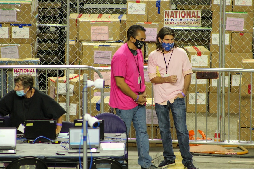 Two men in pink shirts and COVID masks observe people counting election ballots 