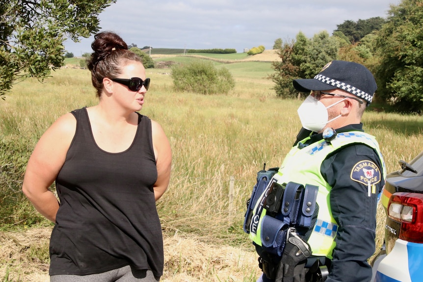 A woman in a black singlet stands talking to a policeman in a rural setting. 