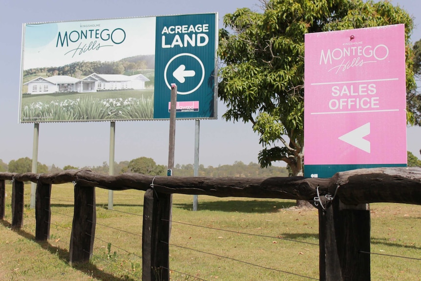 Two signs advertising Montego Hills 'acreage land' and 'sales office' stand on grass behind a timber fence.