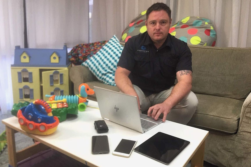 A man sitting on a couch using a laptop and mobile phone, surrounded by children's toys