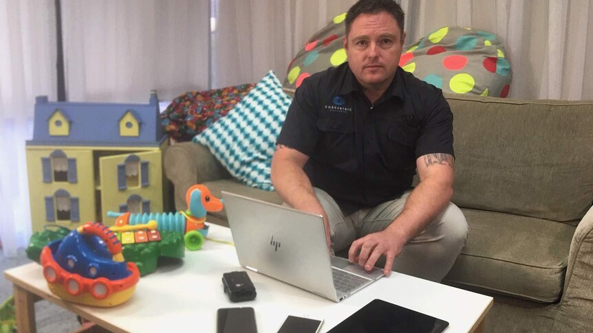 A man sitting on a couch using a laptop and mobile phone, surrounded by children's toys