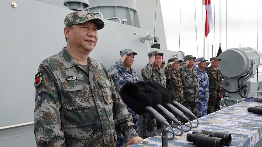 Xi Jinping, in olive, brown and green army fatigues, speaks on a ship after reviewing the naval forces.
