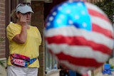 Photo show a middle-aged woman wipes tears from her eye standing on a street in front of a blurred US-flag balloon