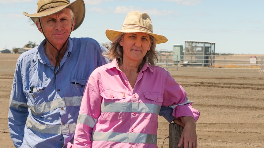 A man and a woman, both wearing hats, stand along a fence on the property boundary with a gas well in the background.