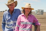 A man and a woman, both wearing hats, stand along a fence on the property boundary with a gas well in the background.