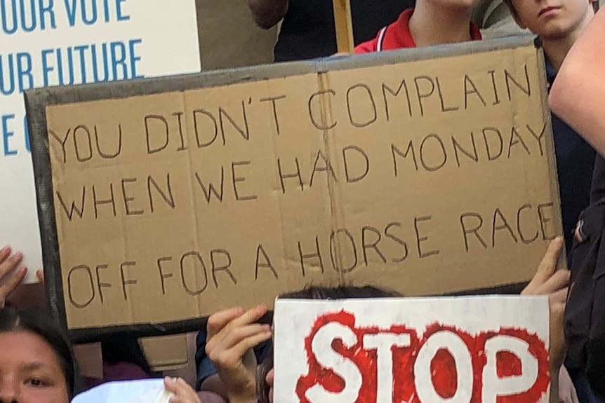 A sign that reads "you didn't complain when we had Monday off for a horse race" is held among a group of children.