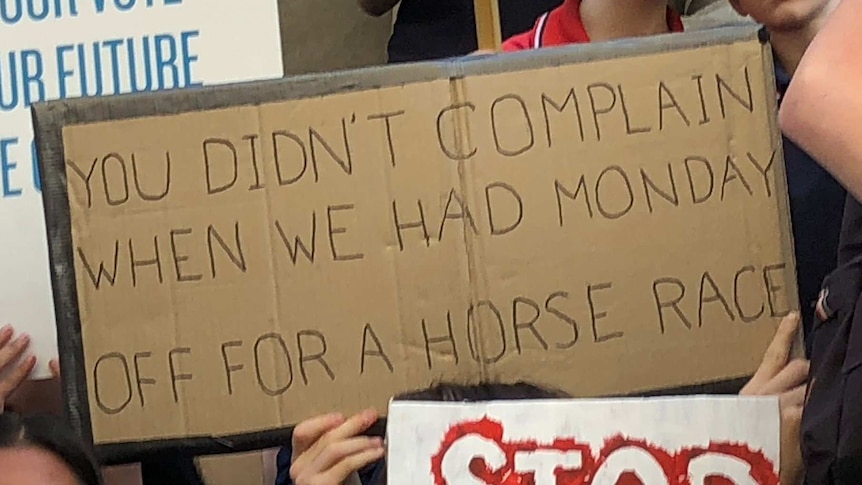 A sign that reads "you didn't complain when we had Monday off for a horse race" is held among a group of children.