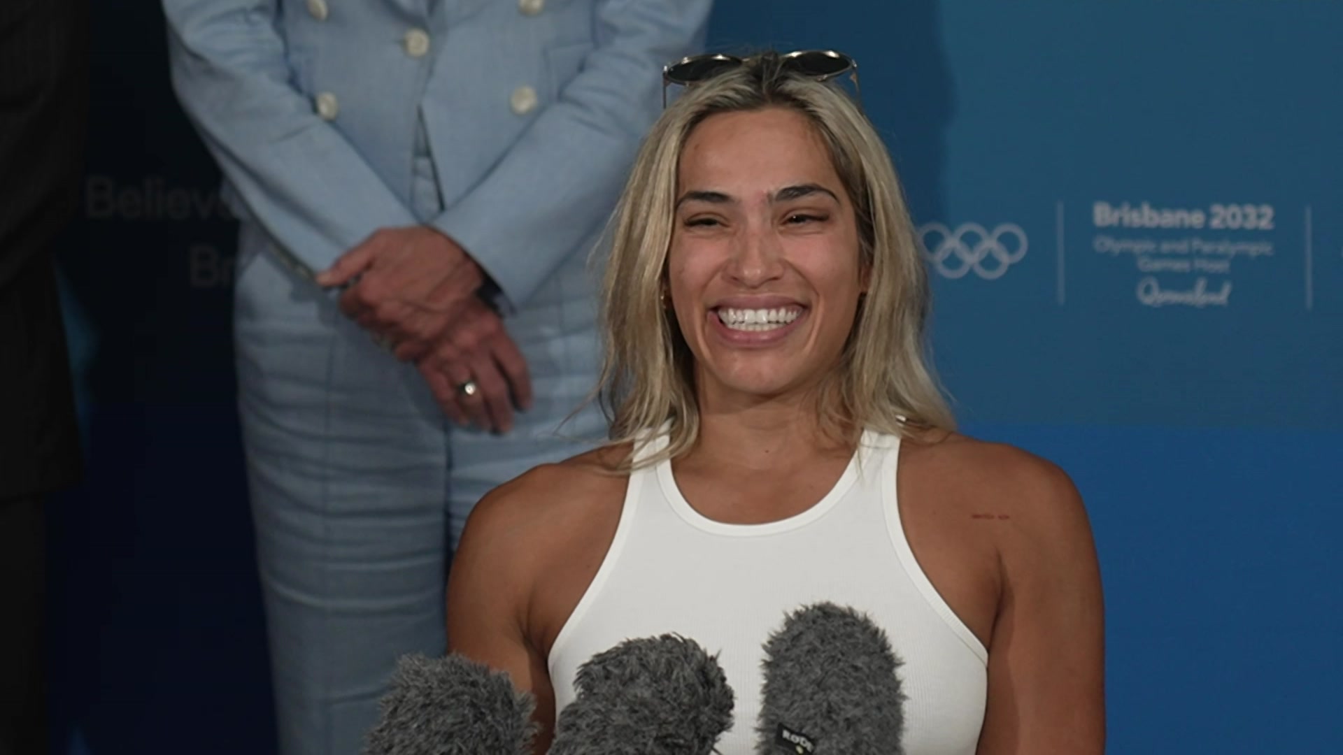 Una mujer rubia con una camiseta blanca y gafas de sol en la cabeza sonríe frente a los micrófonos
