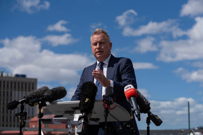 A premier speaks at a press conference on the balcony of a hotel, with the Hobart waterfront in the background.