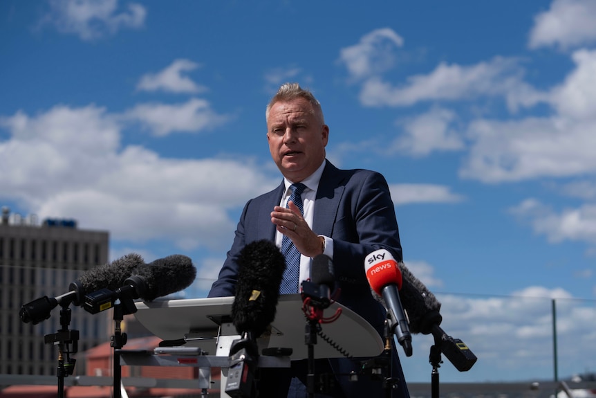 A premier speaks at a press conference on the balcony of a hotel, with the Hobart waterfront in the background.