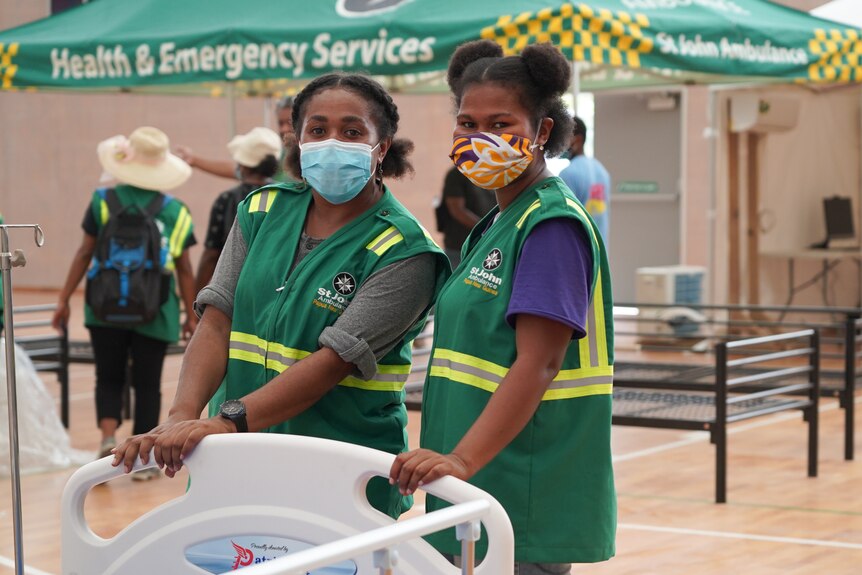 Two women dressed in green uniforms and wearing masks stand over a hospital bed