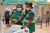 Two women dressed in green uniforms and wearing masks stand over a hospital bed