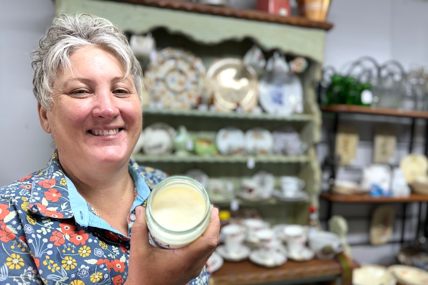 A woman holds up a glass jar of tallow with a blurred shop behind her.