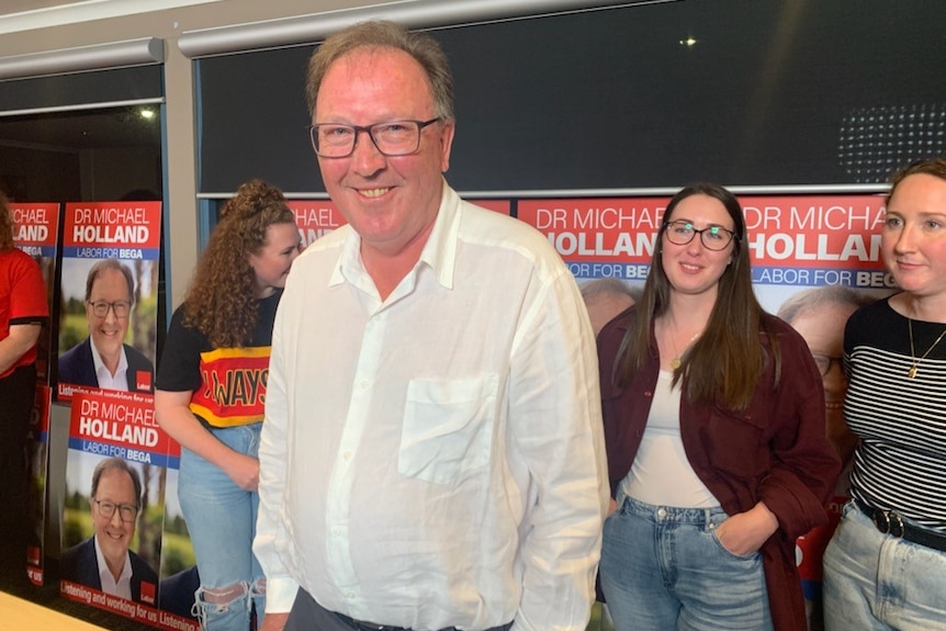 Man in white shirt and glasses, smiling. Several women standing in the background.