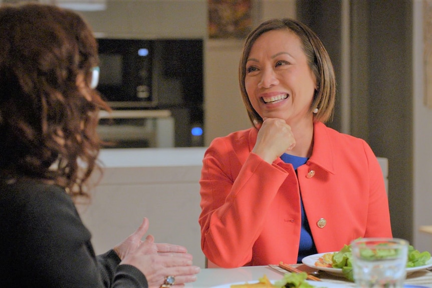 Dai Le wearing a red jacket at a dining table looking and smiling at Annabel Crabb