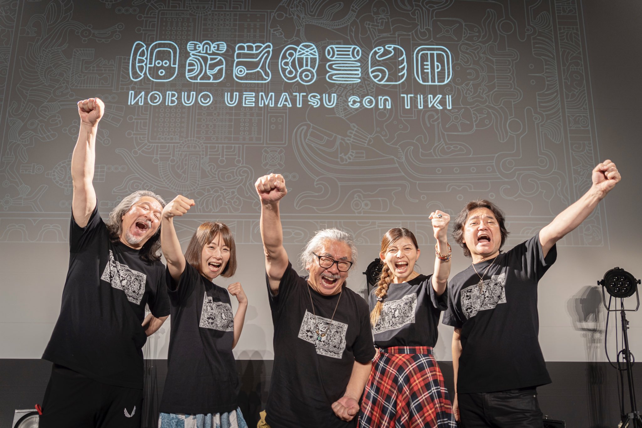 Nobuou Uematsu conTIKI band poses in celebratoin, fists aloft, onstage against screen projection of their band logo