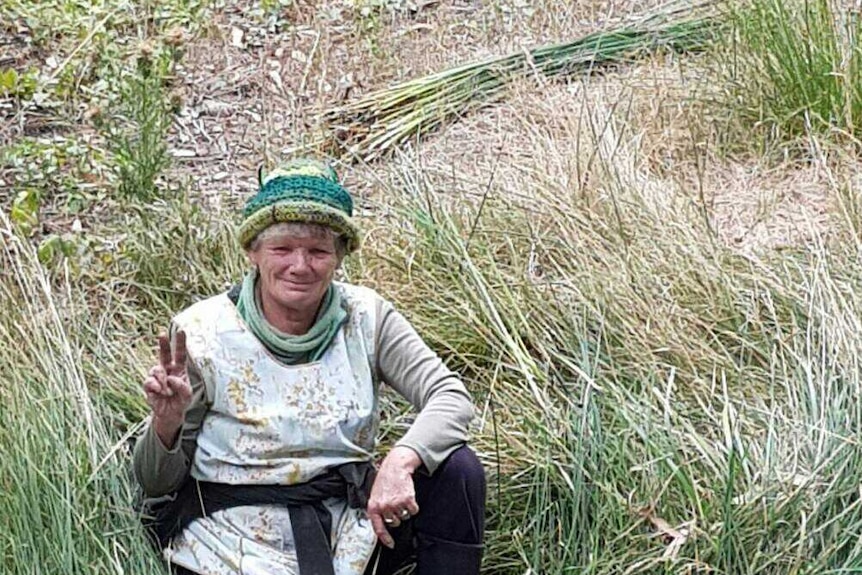 A Tasmania Aboriginal woman sits in long grass smiling and giving a peace sign with two fingers raised.