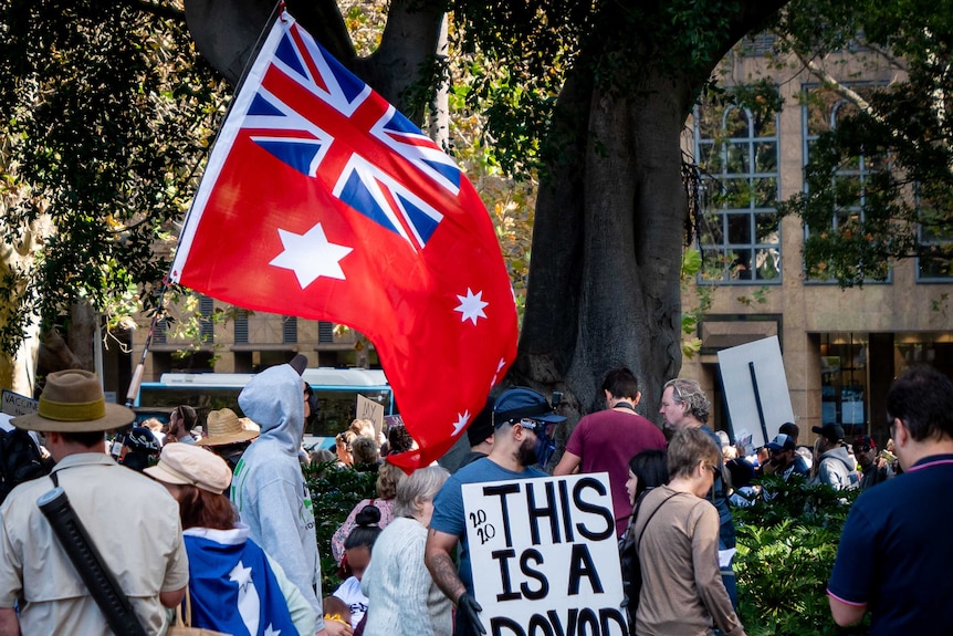 An Australian red ensign flag flies at a coronavirus rally, next to it a man holds a sign that says 'this is a psyop'