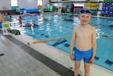 A boy in blue swimmers stands in front of an indoor pool.