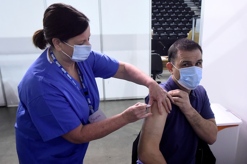 A South Asian man wearing a face mask gets an injection from a female nurse in mask.