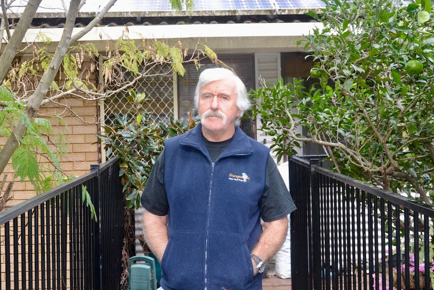 Man standing on balcony with hands in pockets, surrounded by trees.