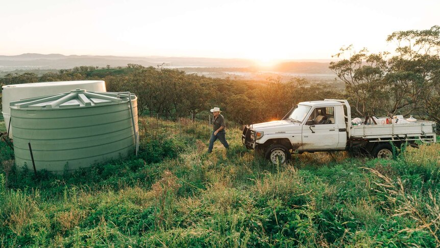 A man with a white hat walking from a ute towards a water tank with a sunrise in the background
