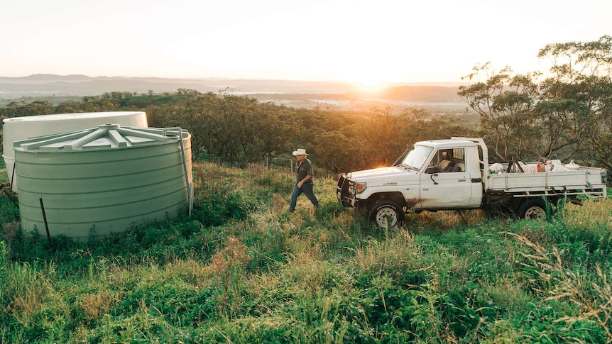 A man with a white hat walking from a ute towards a water tank with a sunrise in the background