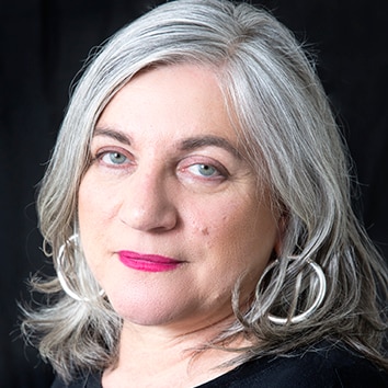 A headshot of a woman with grey hair and pink lipstick and silver hoop earrings  