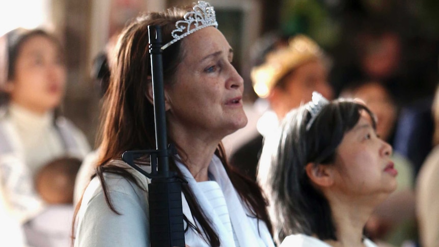 A woman wearing a white gown and tiara holds a rifle at her side while singing