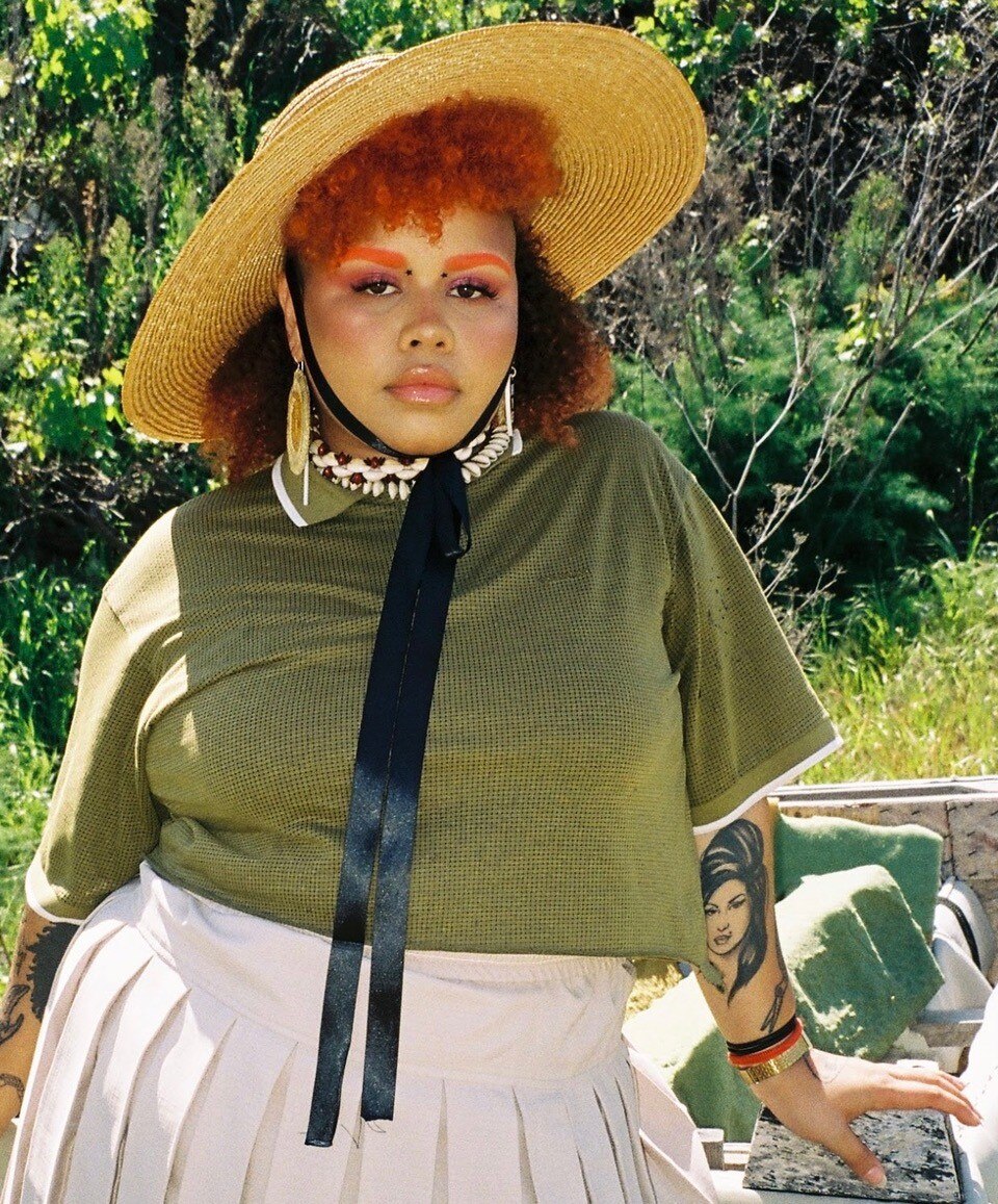 Musician Kaiit stands wearing a straw hat, showing tattoo of Amy Winehouse on her arm