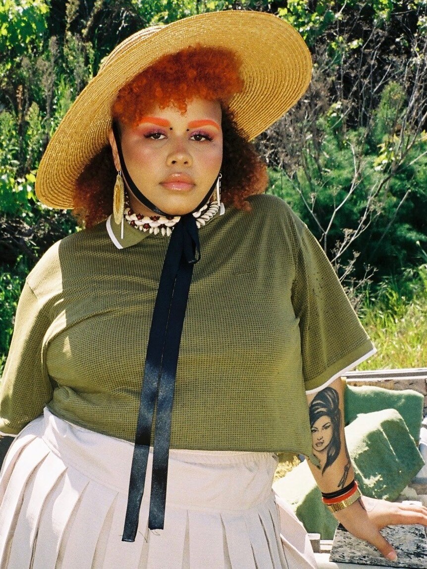 Musician Kaiit stands wearing a straw hat, showing tattoo of Amy Winehouse on her arm