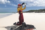 Lifesaver wearing a stinger suit stands on a beach looking for Irukandji jellyfish in water inside a plastic container.