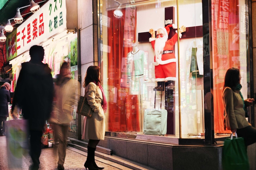 An artwork depicts a tight Japanese street scene where Santa Claus is nailed to a Crucifix and displayed behind a shop window.