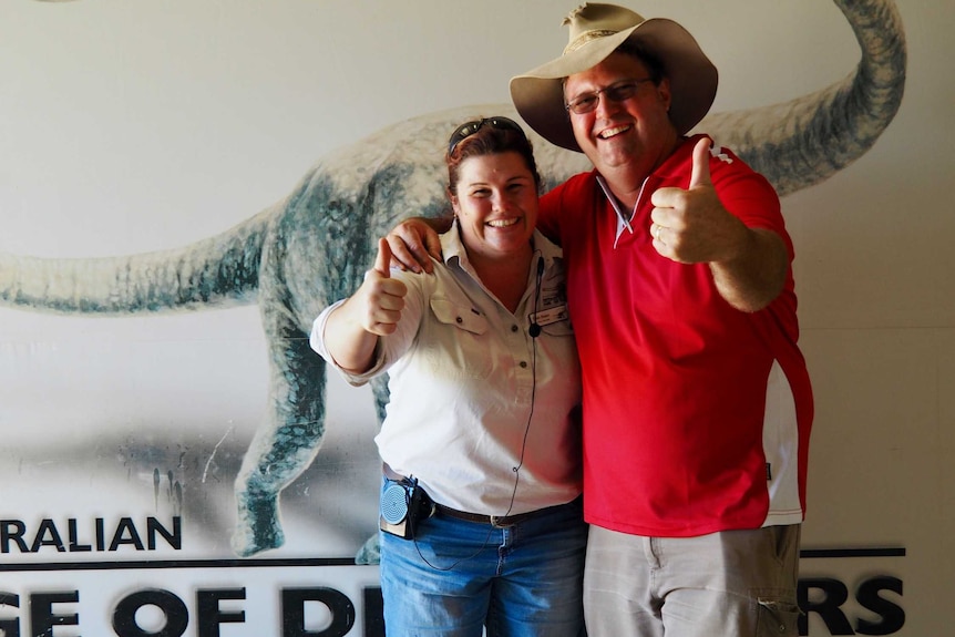 A man and a woman give the thumbs up, standing in front of a dinosaur poster on a wall.