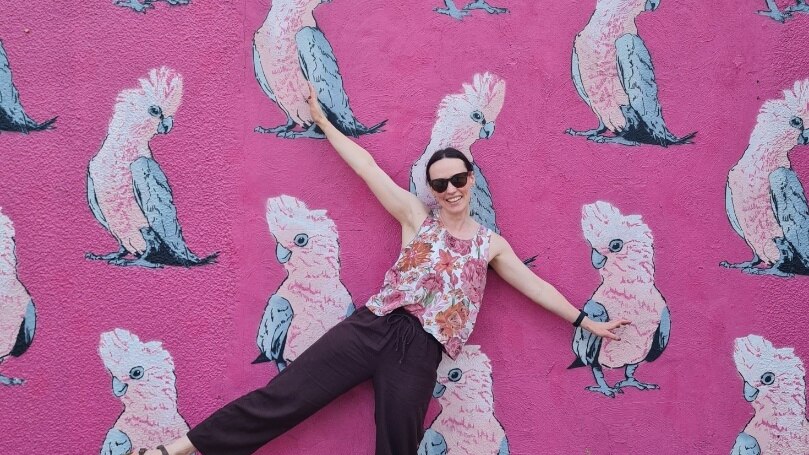 A woman in a pink floral top and black pants standing in front of a pink mural depicting multiple birds